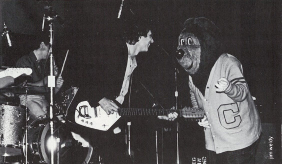 Archival scan: Oski beside a guitarist and drummer onstage. Photo credit: Tim Weldy (printed scan credit).